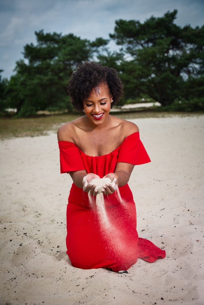 African girl with natural hair and red lipstick playing with sand wearing a red dress